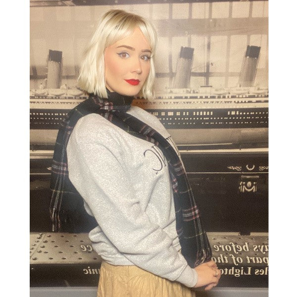 A woman in warm clothes wears a black tartan wool scarf while standing in front of a black and white photo of Titanic.