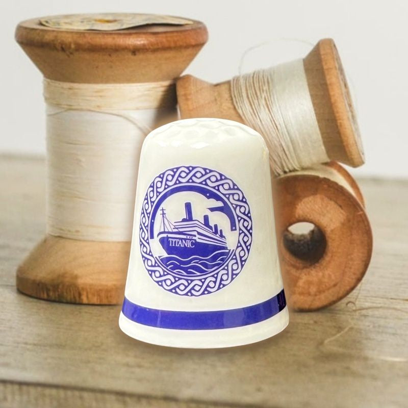 A white ceramic thimble printed with the Titanic in navy blue displayed next to vintage spools of thread.