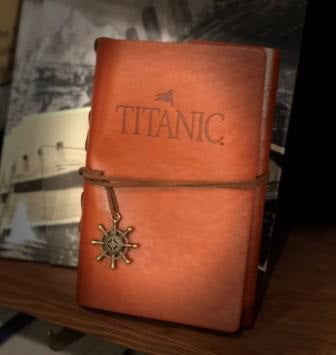 Reddish brown faux leather journal with Titanic's name embossed and a suede strap closure with brassy ship wheel charm.