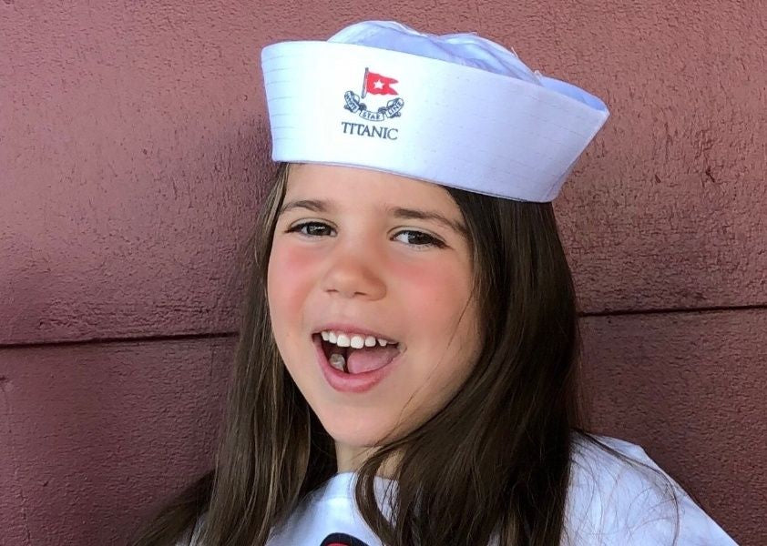 A happy child gives a big grin as they wear a classic white sailor cap printed with the White Star Line logo and Titanic's name on the front.