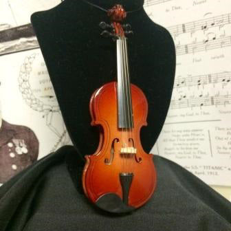 A miniature violin with strings rests against a miniature black velvet bust in front of sheet music. The violin has a black string so it can be used as an ornament.