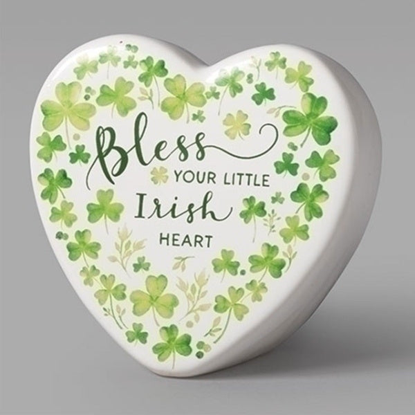 A white ceramic heart decorated with printed green shamrocks. Text in assorted fonts reads "Bless your little Irish heart"