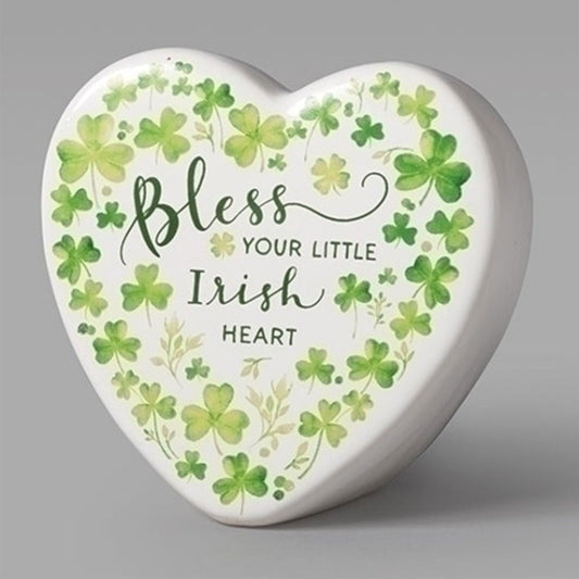 A white ceramic heart decorated with printed green shamrocks. Text in assorted fonts reads "Bless your little Irish heart"