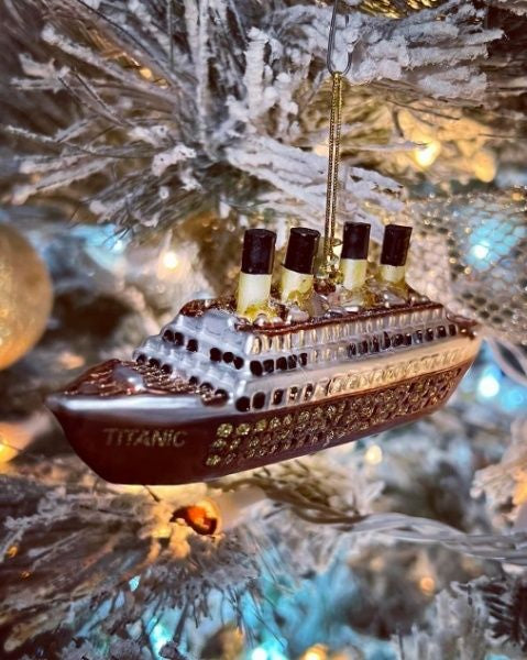 A blown glass Titanic ornament with glittery gold details hangs on a frosty flocked tree.