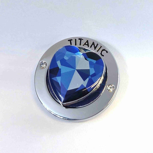 A closeup of a silver-tone shiny round magnet with a printed blue heart spinner piece in the middle. The magnet says Titanic and has two clear gemstones.