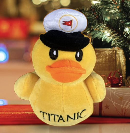A yellow rubber duck style plush toy in a captain hat with embroidery on the tummy that says "Titanic". The toy is sitting under a Christmas tree with presents.