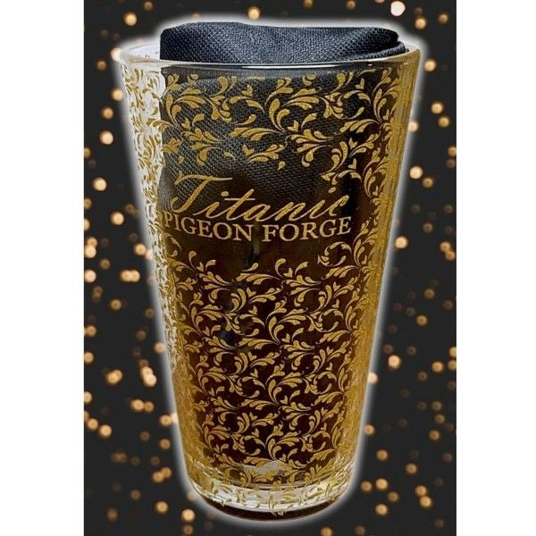 A clear pint glass with an all-over gold filigree print and text that says Titanic Pigeon Forge. The clear glass is displayed with a black cloth inside.