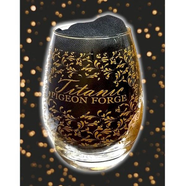 A clear stemless wine goblet with an all-over gold filigree print that says Titanic Pigeon Forge. The clear glass is displayed with a black cloth inside.