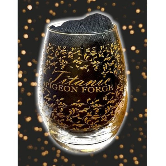 A clear stemless wine goblet with an all-over gold filigree print that says Titanic Pigeon Forge. The clear glass is displayed with a black cloth inside.