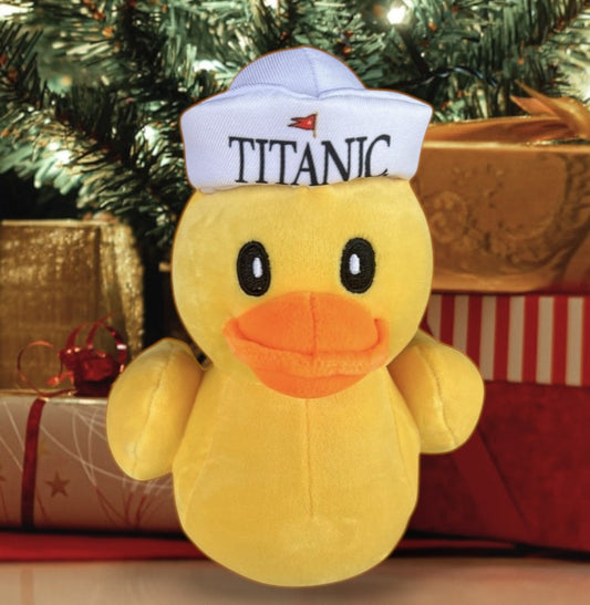 A yellow rubber duck style plush wearing a white sailor hat that says "Titanic". The toy is sitting under a Christmas tree with presents.