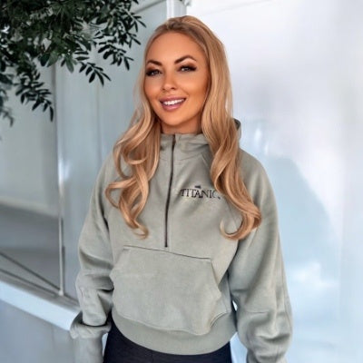 A woman with long hair is wearing a sage green hoodie that zips halfway down. It is cropped at her natural waist and has roomy long sleeves and a kangaroo pocket. A small Titanic logo is printed on one side of the chest.