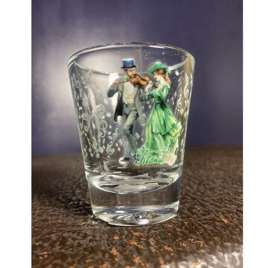 A clear standard shot glass printed with a coupe in formal attire enjoying a cheerful fiddle tune surrounded by pale green music notes.