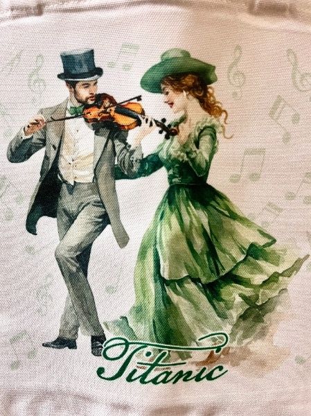 Closeup of the image on the tote bag. A man in a suit is playing the fiddle while a woman with red hair in a green gown enjoys the tune. They are surrounded by pale green music notes, and the name Titanic is at their feet.