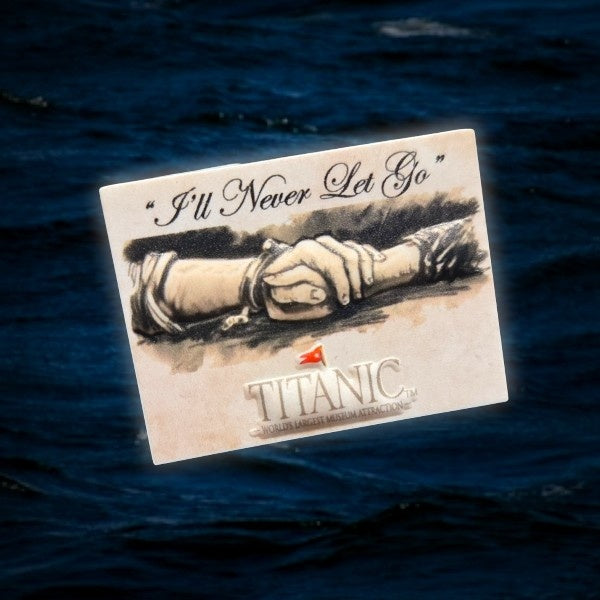 A miniature rectangular magnet, about the size of a USD quarter, with the words "'ll Never Let Go" above a sketch illustration of two clasped hands. Our Titanic logo is printed below. Staged on a photo of dark blue ocean water.
