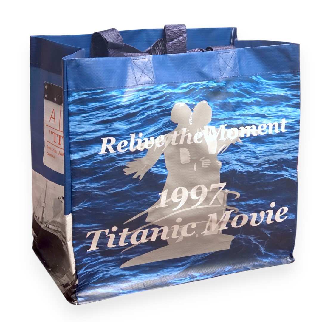 Blue shopping tote with a greyscale silhouette of Jack and Rose on the bow of the Titanic against a background of blue waves. Large white text says "Relive the Moment, 1997, Titanic Movie".