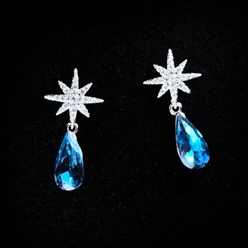 A pair of white post starburst earrings with a dangling blue teardrop gem.