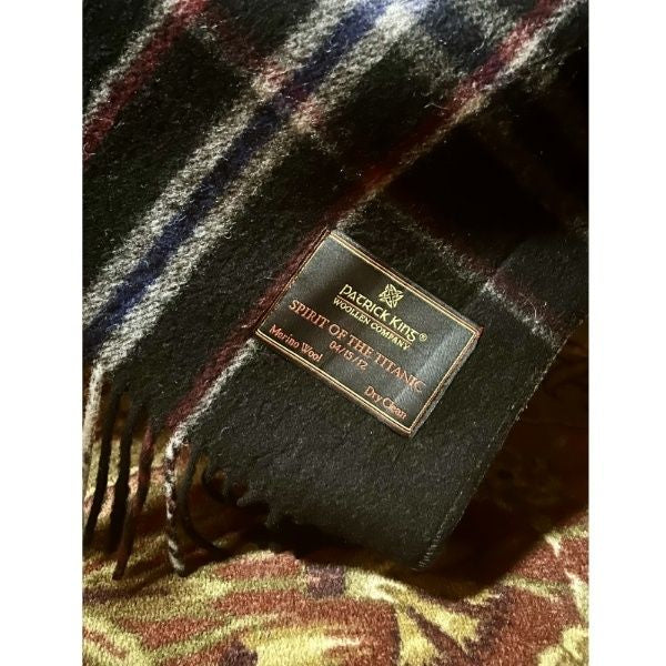A closeup of the embroidered black tag on the corner of  black plaid scarf. It says Patrick King Woollen Company, Spirit of the Titanic, 04.15.12, Merino Wool, Dry Clean.