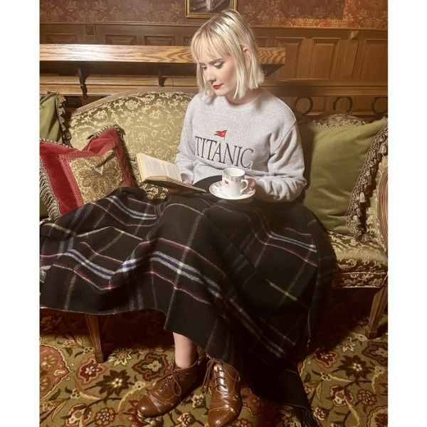 A young woman sits on a couch with a black plaid knee blanket across her lap. The blanket is wide enough to wrap up in or share, but narrow enough that it won't pool on the floor.