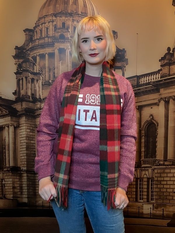 A young woman models a red and green plaid scarf draped loose over her shoulders, the fringe reaches her hips.