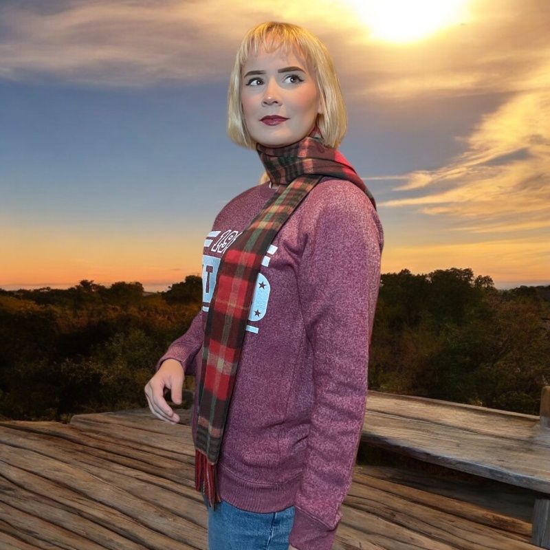 A young woman models a red and green plaid scarf, worn around her neck. She is standing in front of an autumnal sunset.