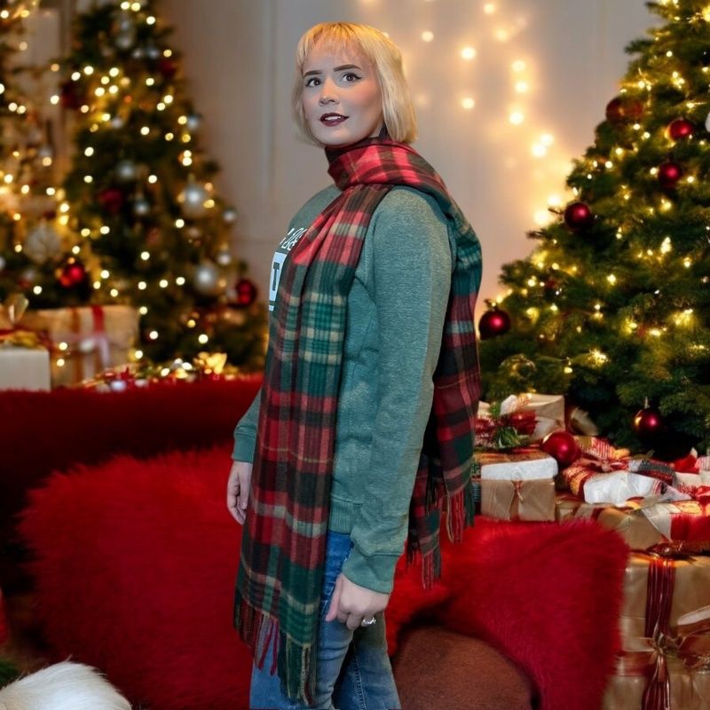 A blonde young woman models a red and green plaid stole as if it is a large scarf. She is wearing complimenting green and standing near Christmas trees.