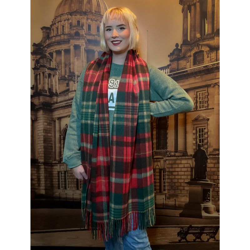 A blonde young woman models a red and green plaid stole draped around her shoulders, the fringe of the stole nearly reaches her knees.