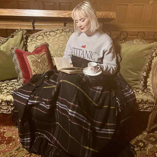 A young woman is reading on a vintage couch with a cup of tea, comfortably wrapped up in a deluxe wool blanket in plaid colors of black, white, red, blue, and gold.