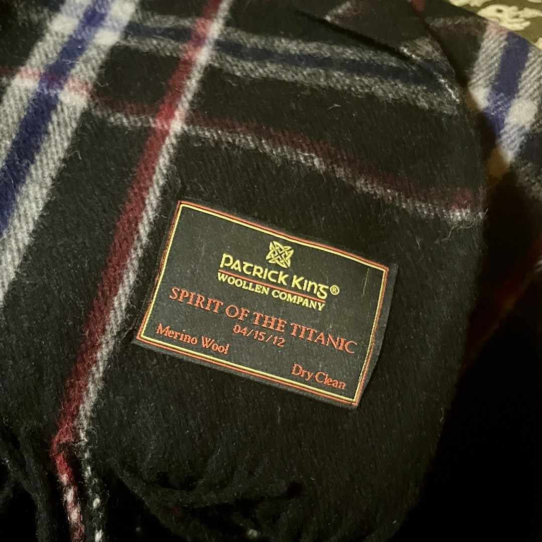 A closeup of the embroidered tag on the wool blanket, the blanket's fringe can be seen on one corner. The tag says Patrick King Woollen Company, Spirit of the Titanic, 04/15/12, Merino Wool, Dry Clean.