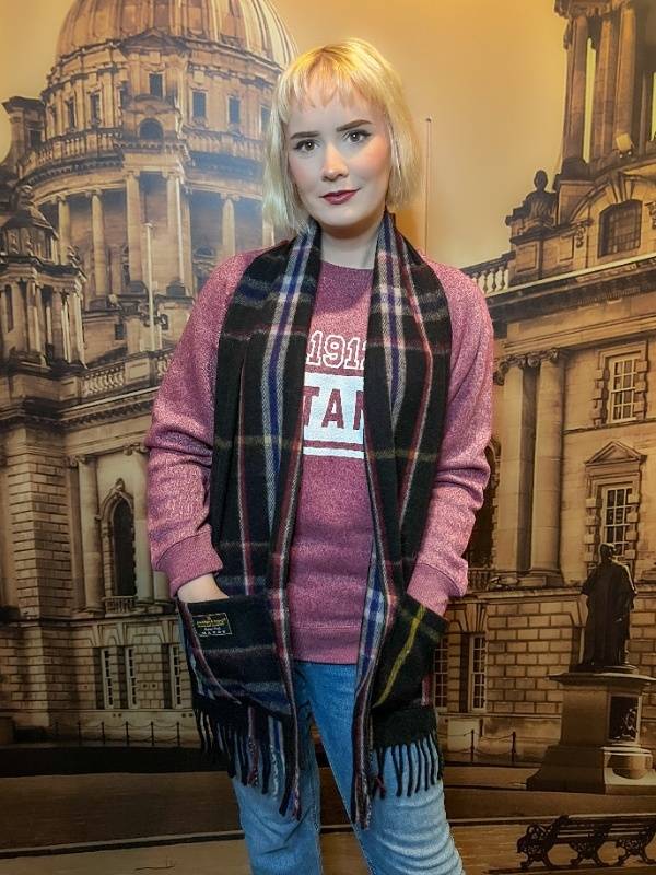 The same blonde woman wears a different scarf from the same Spirit of Titanic line, displaying that the placement of the stripes in the knitting varies with each scarf. This black scarf features navy stripes more prominently than gold.