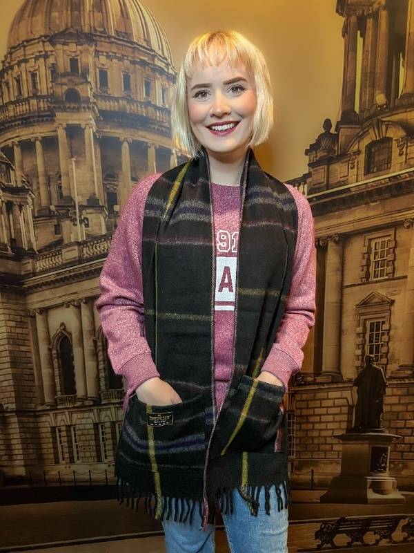 A blonde woman smiles as if to say "It has pockets!" while wearing this Titanic scarf. The scarf is black with subtle grey, navy, red, and gold stripes.