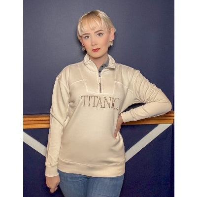 A woman wearing a pale beige pullover sweatshirt with a quarter zip lapel and embroidered with large beige letters across the chest that say Titanic.