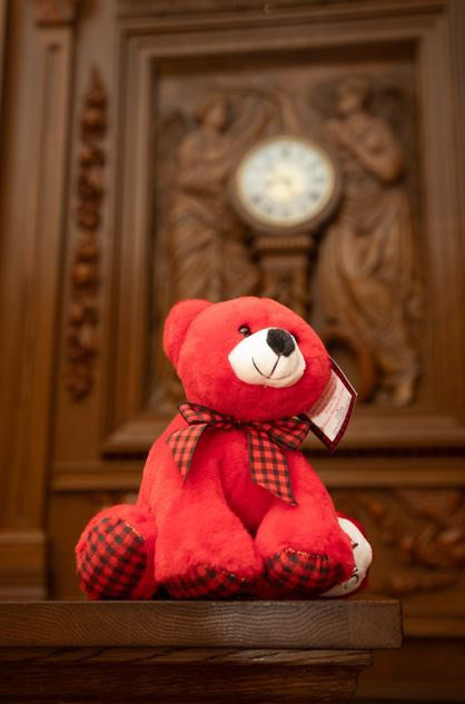 A small red bear with plaid paws and matching plaid bow is sitting in front of the Titanic's grand staircase clock. The red bear has a white snout and black nose, and one paw is white with a Titanic logo embroidered.