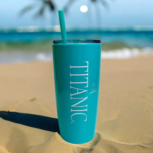 A bright blue-green tumbler with matching straw sits on a sandy beach, it has a large print of our Titanic logo in white down the side.