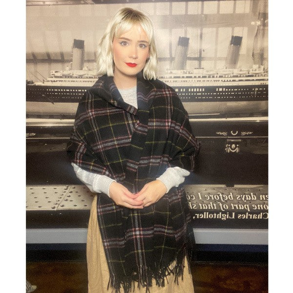 A woman has a black wool tartan stole wrapped around her shoulders and arms as she stands by a photo of the Titanic.