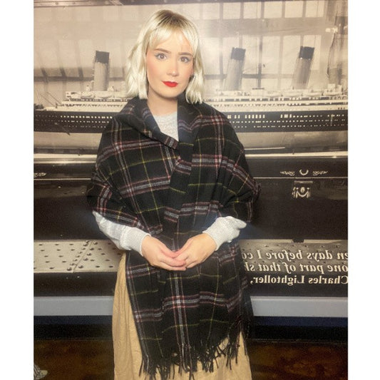 A woman has a black wool tartan stole wrapped around her shoulders and arms as she stands by a photo of the Titanic.