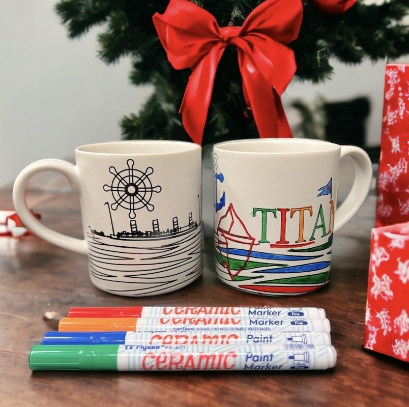 One uncolored and one colored customizable mug with a Titanic theme and ceramic paint markers. The product is on a festive Christmas table.