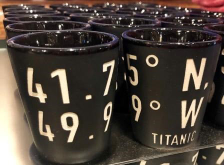 Matte black shot glasses sit on a shelf, they are engraved with bold white numbers and the name Titanic.