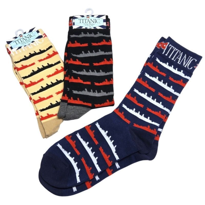 All three colors of Titanic ship pattern socks, tan, black, and navy. The tan and black are folded with their tag still on, and the navy socks are unfolded. Ship silhouettes repeat in alternating colors across the entire sock and Titanic's name is on the opening at the top.
