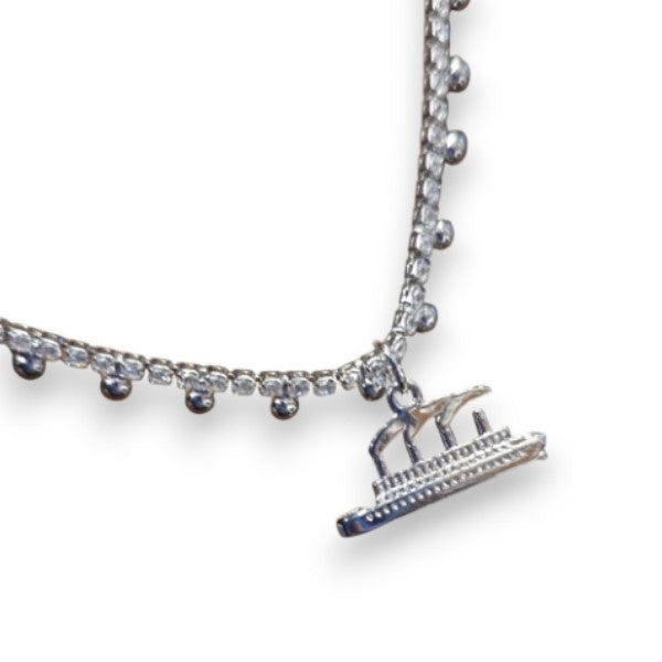 A closeup of a silver-tone necklace with a rhinestone chain that has little silver dot accents, and a silvery Titanic charm with smoke coming from 3 smokestacks. It is dainty, and delicate.