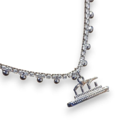 A closeup of a silver-tone necklace with a rhinestone chain that has little silver dot accents, and a silvery Titanic charm with smoke coming from 3 smokestacks. It is dainty, and delicate.