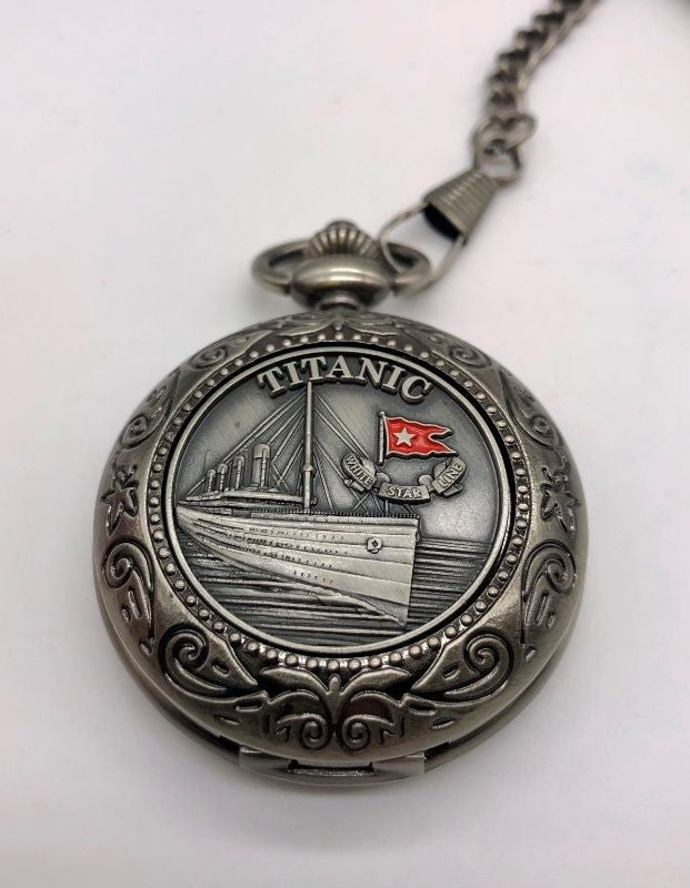 A dark silver-tone antique style pocket watch with swirling design on border and pewter emblem of the Titanic with a small red enamel nautical flag detail.